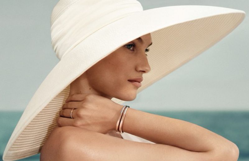 Model Greta Hofer wearing an oversized cream wide-brim sun hat and metallic bangles against a calm ocean horizon.