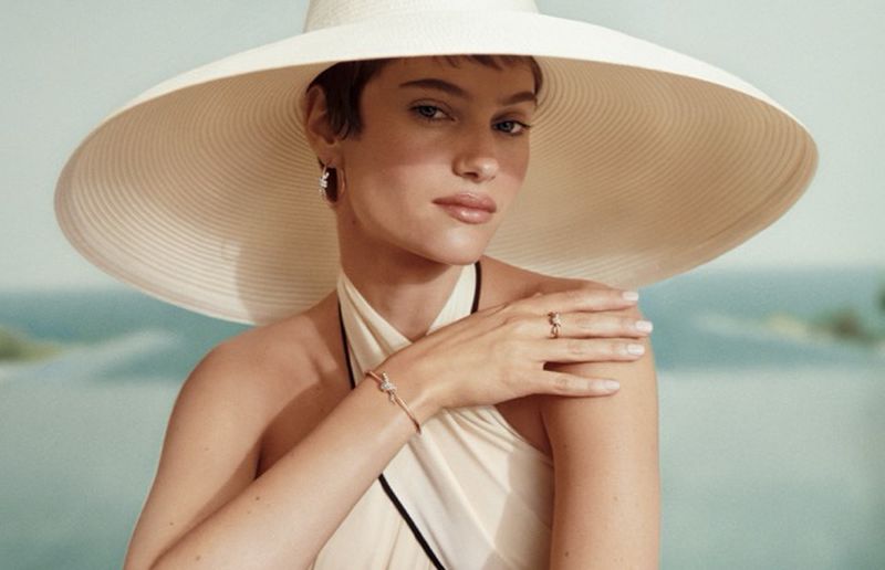 Greta Hofer wearing an oversized cream wide-brim sun hat and fine jewelry against a coastal ocean backdrop.