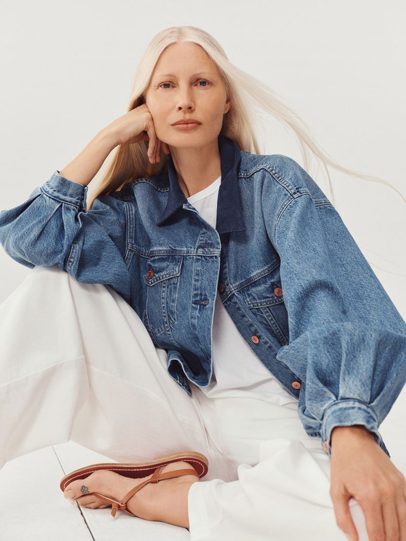 Attractive woman with long white hair is sitting, wearing a denim jacket and white pants in a fashion-style studio shot.
