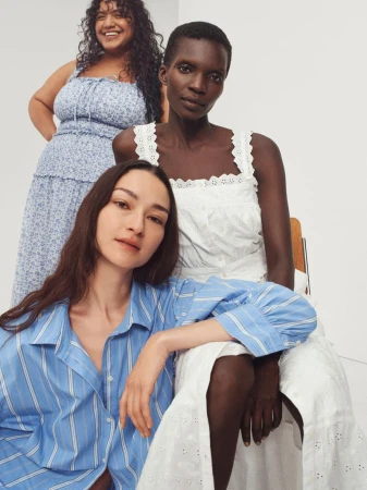 Three women pose indoors in summer dresses: one in a blue floral dress, one in a white eyelet dress, and one in a blue striped shirt.
