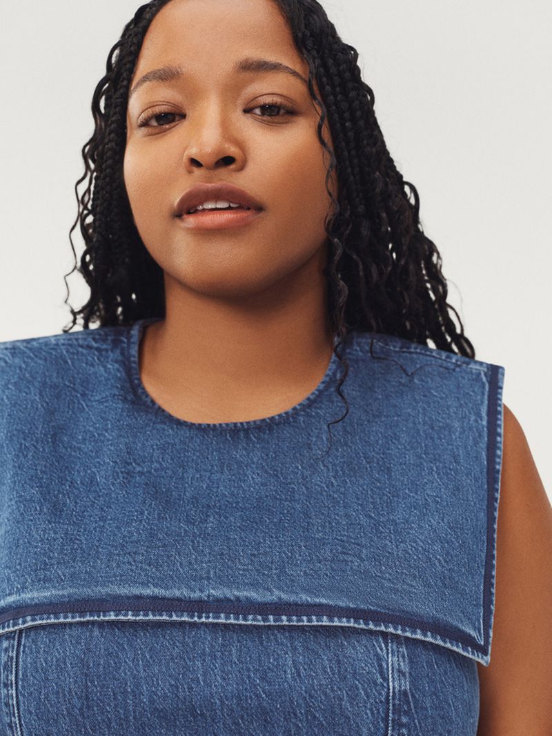Close-up studio portrait of a Black female model in a denim top with braided hair, neutral expression, and simple white backdrop.