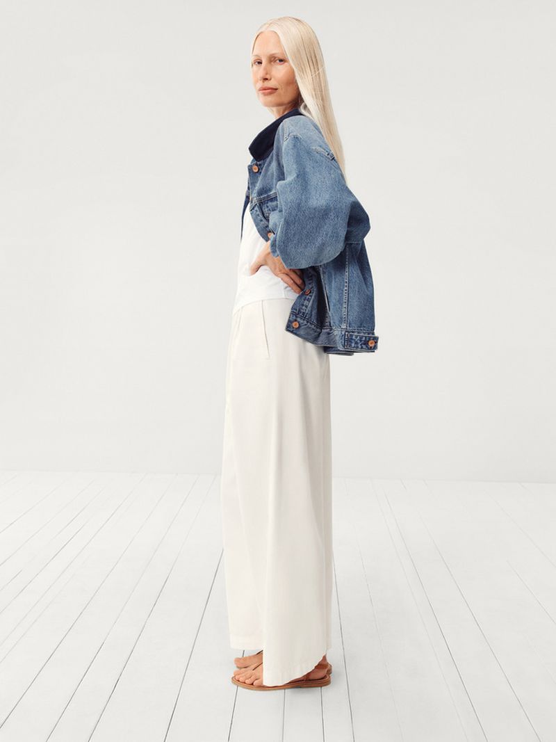 Full shot of a tall, fair-skinned, mature woman in a denim jacket and white palazzo pants standing in a white studio setting, hands on hips.