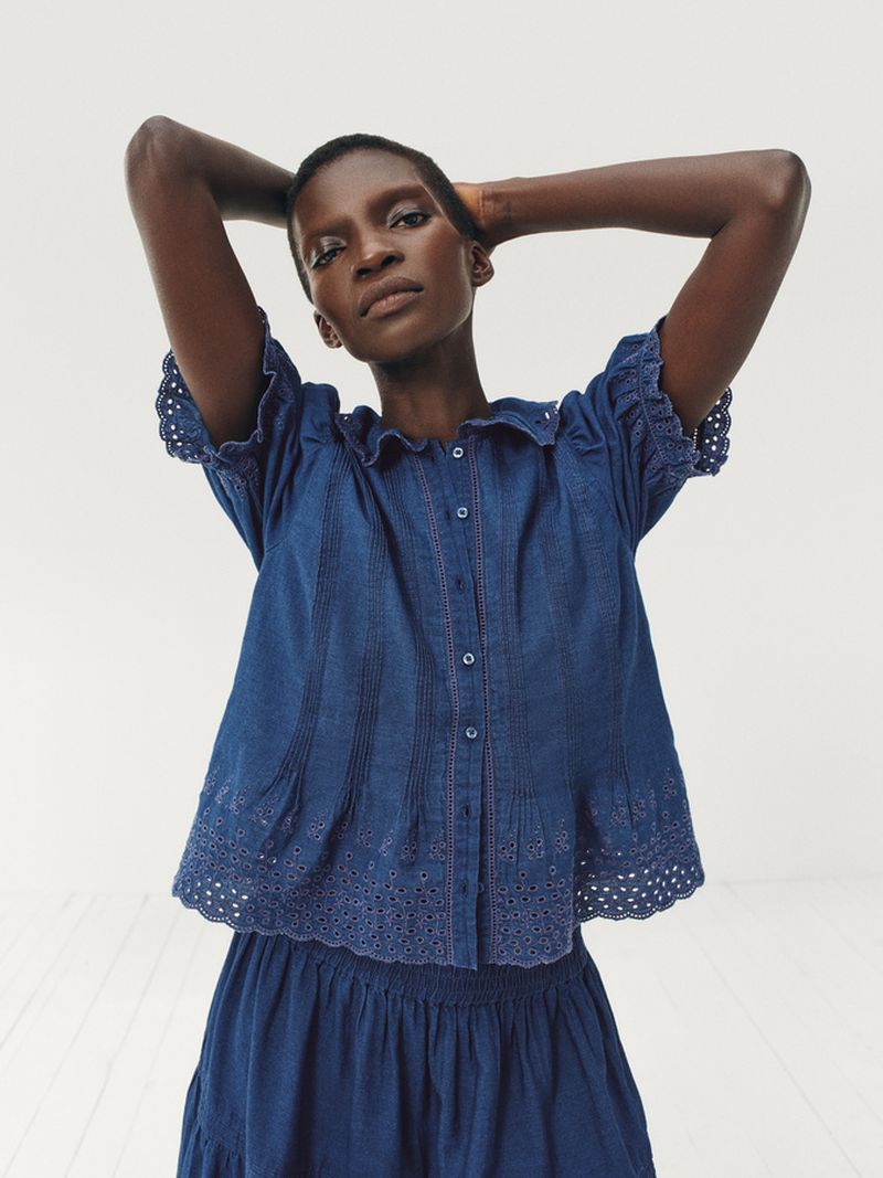 A dark-skinned woman with short hair poses with her arms up wearing a blue denim top and skirt against a neutral backdrop indoors.