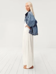 Model Kirsty Hume posing in profile wearing a denim jacket and wide-leg white trousers in a white studio.