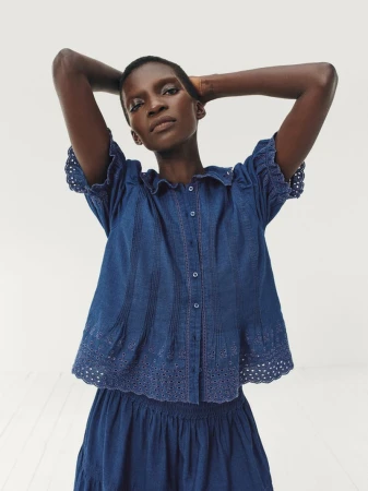 Model Achok wearing a blue eyelet denim blouse and matching skirt with hands behind her head against a white background.
