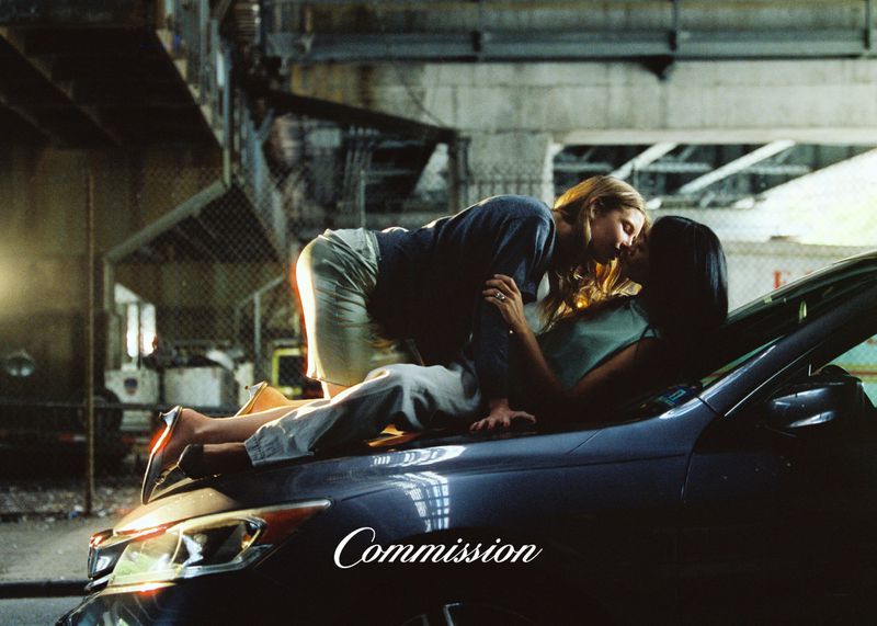 Two women lying intimately on the hood of a dark car beneath an urban overpass for the Commission SS26 campaign.
