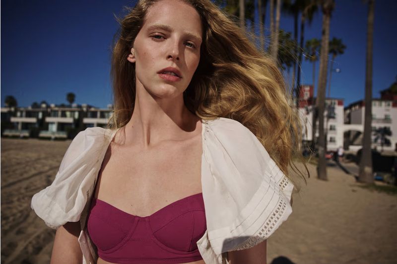 Model Abby Champion wearing a magenta bustier top and white cropped overlay at a sunny beach with palm trees.