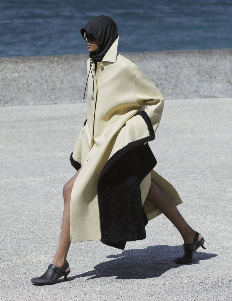 Model Livia Eduarda strides outdoors wearing an oversized pale yellow coat with black lining, a black balaclava, and sunglasses.