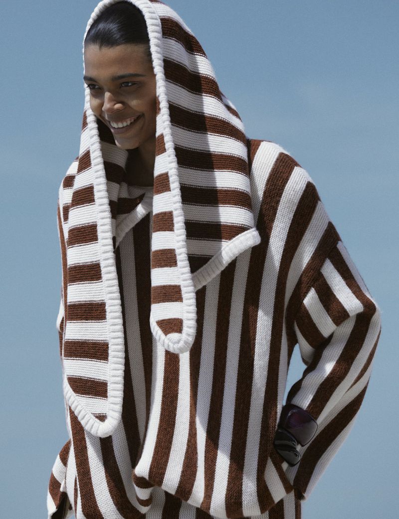 Model Livia Eduarda smiling outdoors wearing an oversized brown and white striped hooded knit sweater under a clear blue sky.