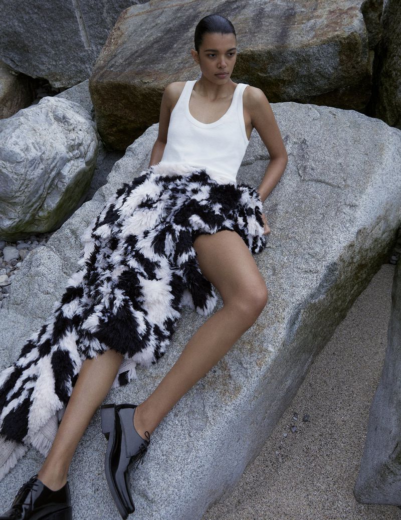 Model Livia Eduarda wearing a white tank top, fuzzy monochrome skirt, and black patent shoes reclined on granite rocks.