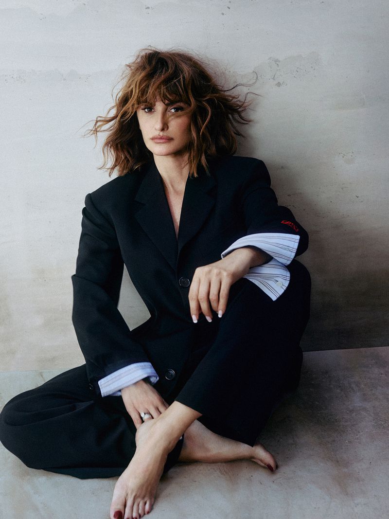 Penelope Cruz barefoot in an oversized black suit sitting against a neutral textured wall for Porter Magazine.