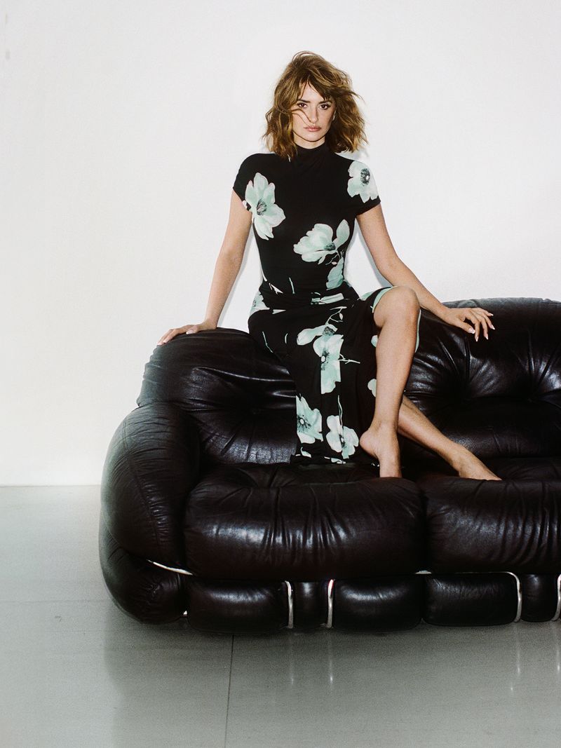 Penélope Cruz wearing a black floral print dress seated barefoot on the back of a black tufted leather sofa against a white wall.
