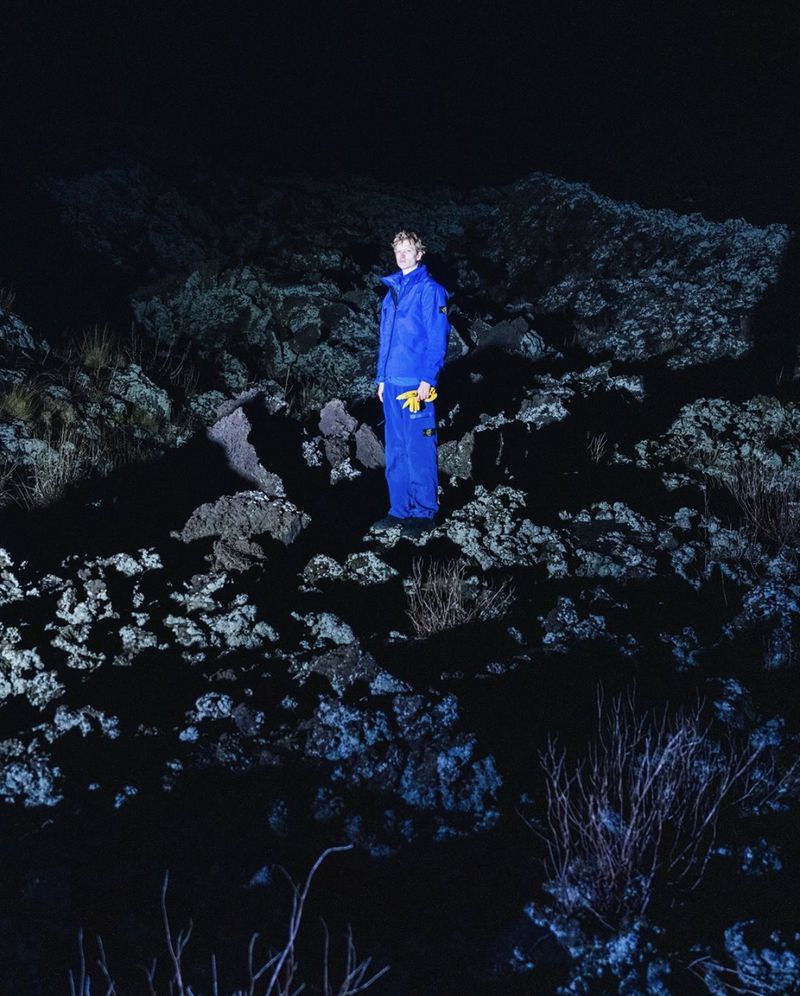 Model wearing a vibrant blue technical suit and yellow gloves standing on rocky terrain at night under direct flash.