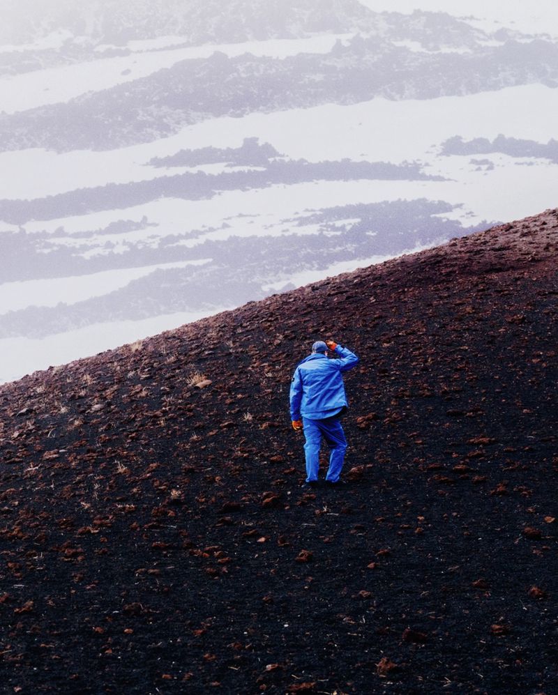 A lone person in a blue jacket and blue trousers walking up a dark volcanic mountain slope under an overcast sky.