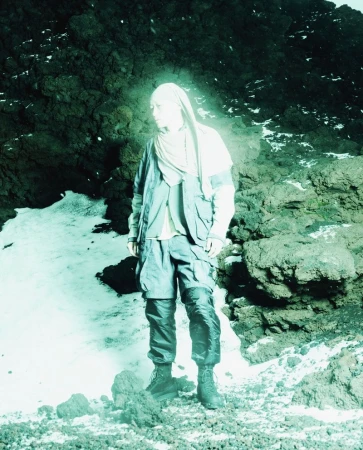 Model wearing layered technical outerwear with a draped headscarf standing on rocky, snow-dusted terrain with a cyan glow.
