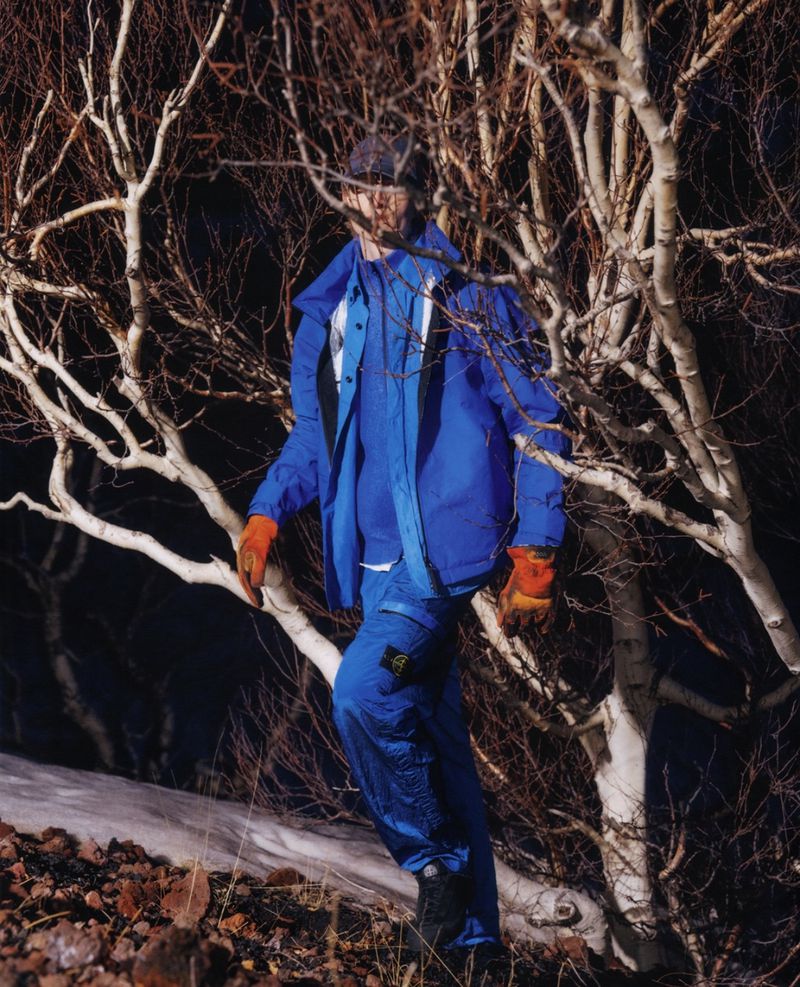 Model wearing a royal blue hooded technical jacket, matching cargo trousers, and orange work gloves amidst bare birch trees at night.