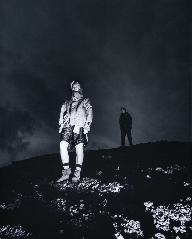 Low angle shot of a model in a hooded technical jacket and shorts standing on a rocky hillside at night with another model behind.