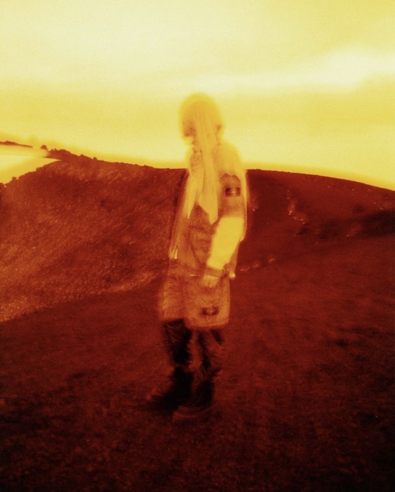 A stylized figure in technical outerwear stands on a dark rugged ridge under an intense glowing yellow sky.