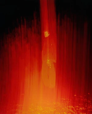 A figure standing enveloped in vertical red light motion blur against a dark background.