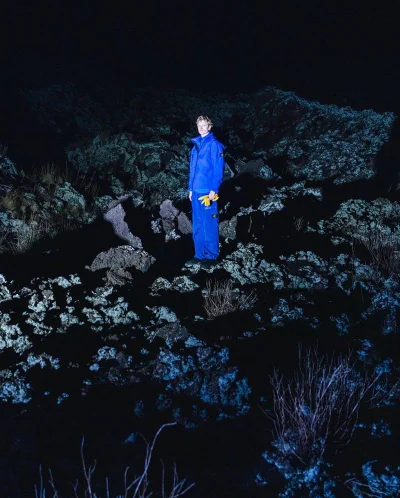 Model wearing a vibrant blue technical suit and yellow gloves standing on rocky terrain at night under direct flash.