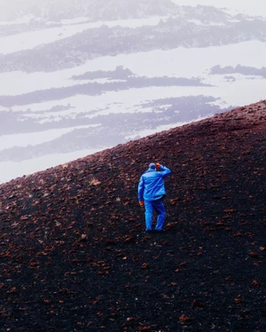 A lone person in a blue jacket and blue trousers walking up a dark volcanic mountain slope under an overcast sky.