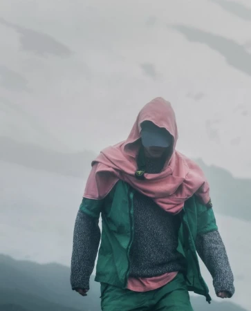 Figure walking outdoors in a green technical jacket, pink hooded wrap, grey knit sweater, and baseball cap against a misty mountain background.
