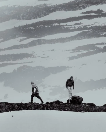Two figures in hooded outdoor apparel walking along a dark rocky ridgeline against a snowy mountain slope.
