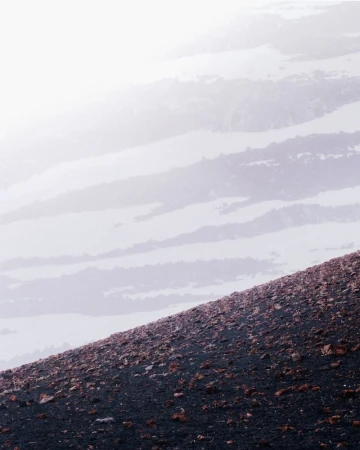 Steep volcanic gravel slope dotted with reddish rocks overlooking mist-shrouded mountain ridges fading into a bright white fog.