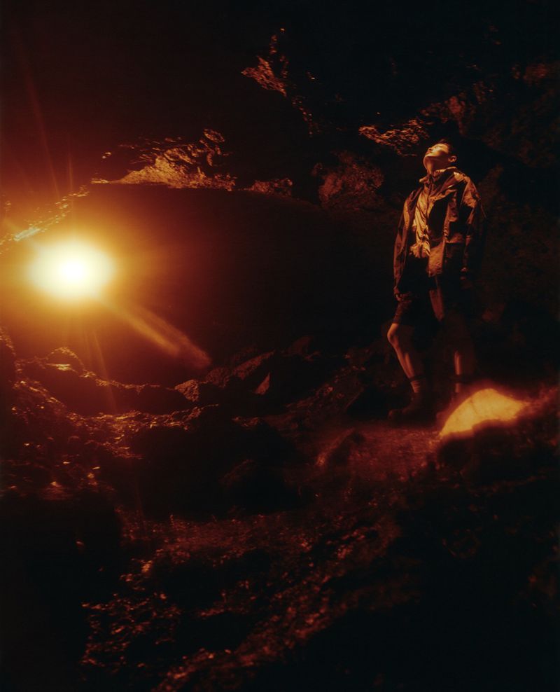 Model looking upwards in a dark rocky cave illuminated by a bright warm lens flare from the side.