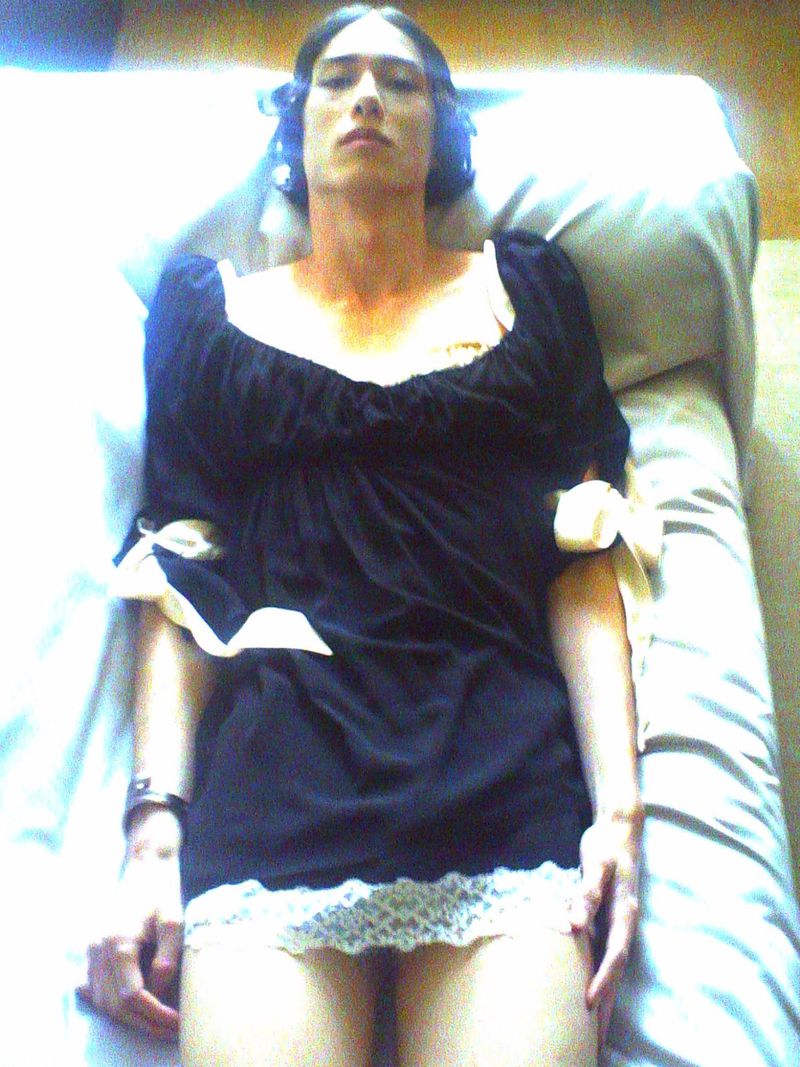 Model lying on a bed wearing a short black dress with white lace trim and ribbon bow accents on the sleeves.