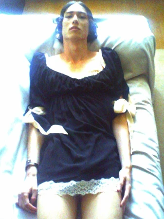 Model lying on a bed wearing a short black dress with white lace trim and ribbon bow accents on the sleeves.