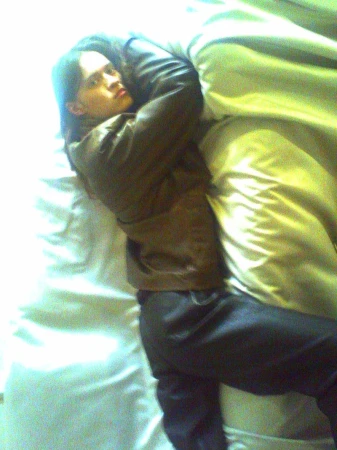 A person in a brown leather jacket and dark trousers lies on white bedding while clutching a pillow.