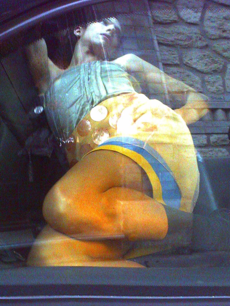 Model reclining inside a vehicle seen through a reflective car window wearing a yellow embellished mini skirt and structured top.