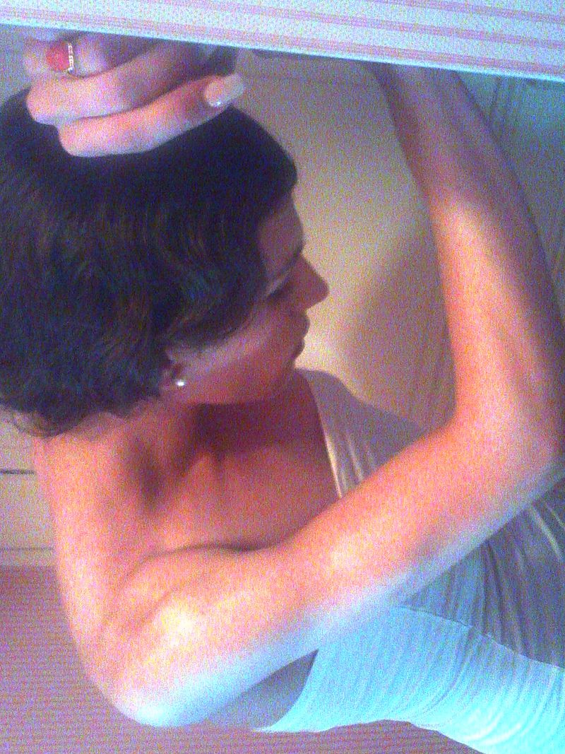 Model with short dark wavy hair wearing a white strapless top with arm raised overhead in an interior setting.