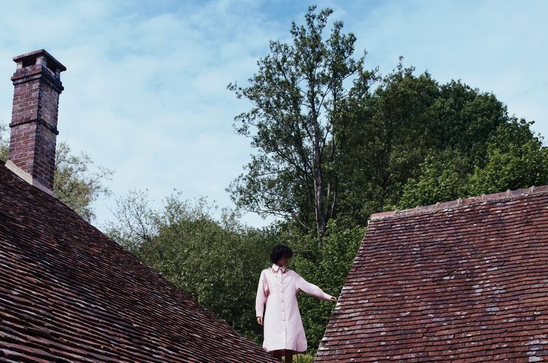Model in a pale pink coat dress standing outdoors between tiled house rooftops against lush green trees.