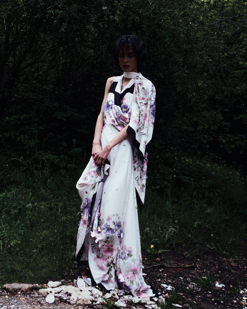 Model Gabrielle Rul standing outdoors wearing a draped floral white dress against dark forest foliage.