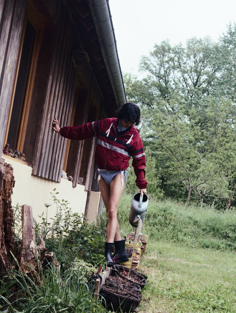 Model Gabrielle Rul pouring a watering can beside a wooden house exterior in a burgundy windbreaker and briefs.