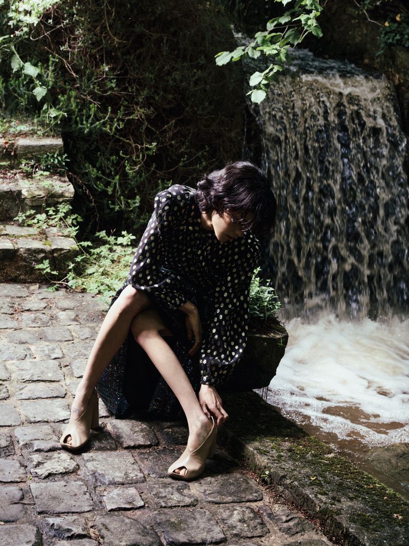 Model Gabrielle Rul in a black polka dot dress sitting on stone steps by a rushing woodland waterfall.