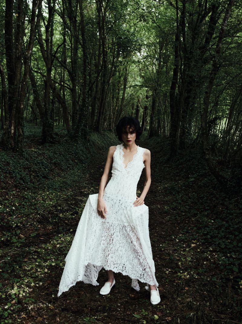 Model Gabrielle Rul in a sleeveless white lace maxi dress posing on a shaded woodland path surrounded by dense forest trees.