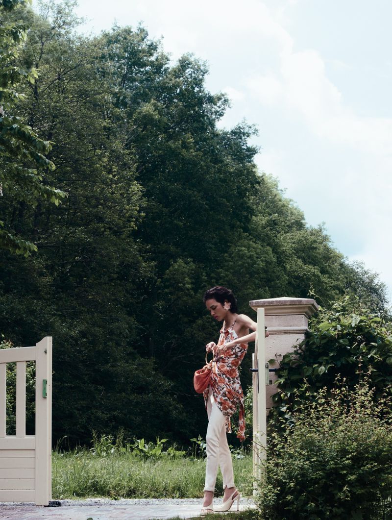 Model Gabrielle Rul wearing a printed halter top, white trousers, and heels standing by a stone gate post outdoors.