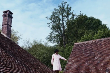 Model in a pale pink coat dress standing outdoors between tiled house rooftops against lush green trees.