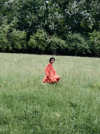 Model Gabrielle Rul in a bright coral flowing dress standing in a tall grass field framed by trees.