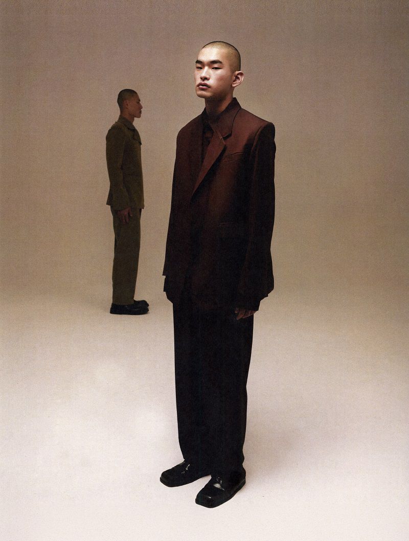 An East Asian male model with a buzz cut poses in a dark brown oversized suit in front of another model on a beige seamless backdrop.