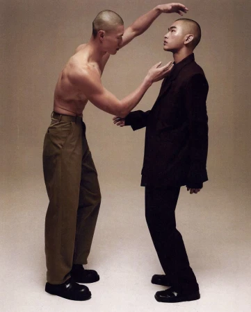 Two male models with shaved heads posing together in a studio, one shirtless in khaki trousers and one in an oversized dark suit.