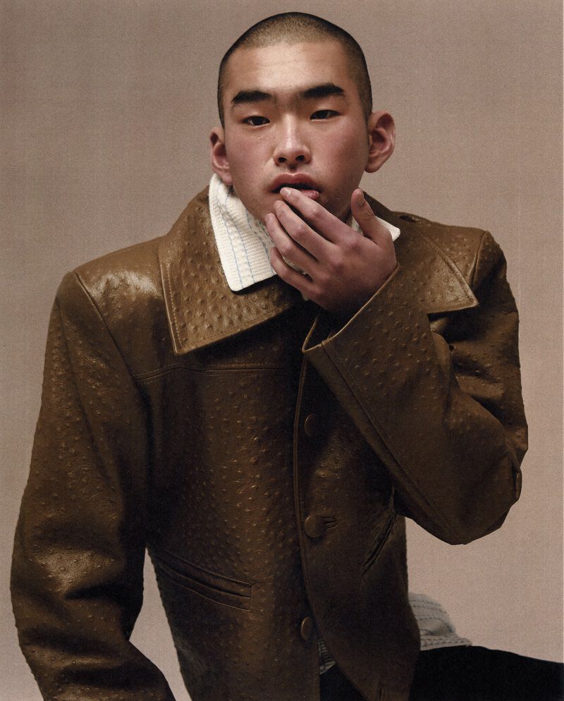 East Asian man with a buzz cut wearing an olive textured leather jacket and white knit collar with fingers touching his lip.