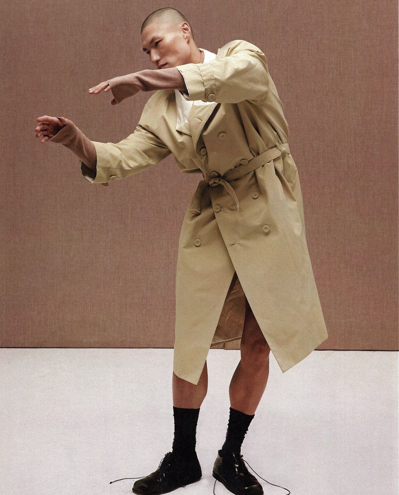 Man wearing a beige trench coat and black shoes posing against a textured brown studio background.