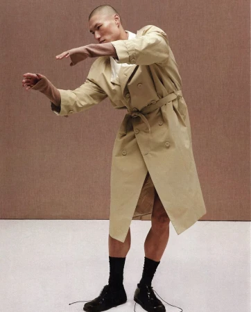 Man wearing a beige trench coat and black shoes posing against a textured brown studio background.