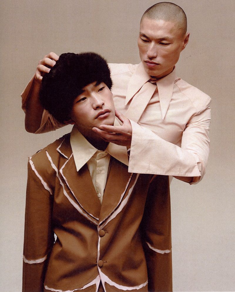 Two East Asian male models in tailored apparel, one adjusting the head and chin of the other wearing a fur hat and brown suit.