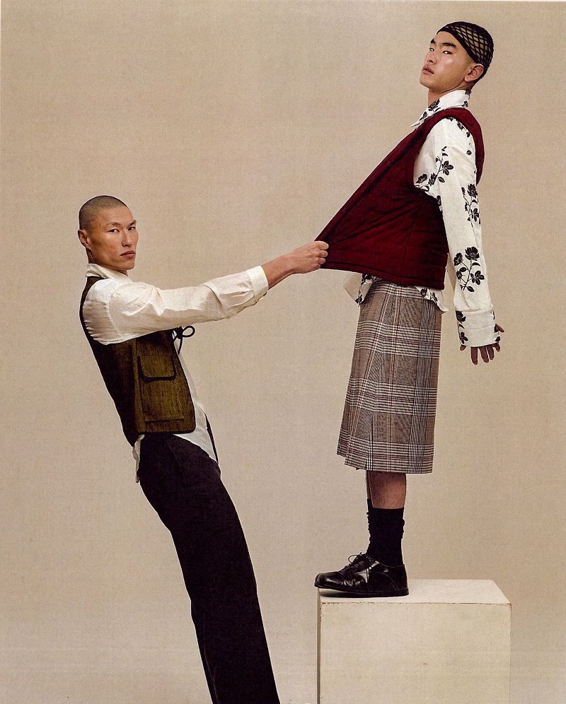 Two men in layered designer menswear and a plaid pleated skirt posing dynamically against a warm neutral studio backdrop.