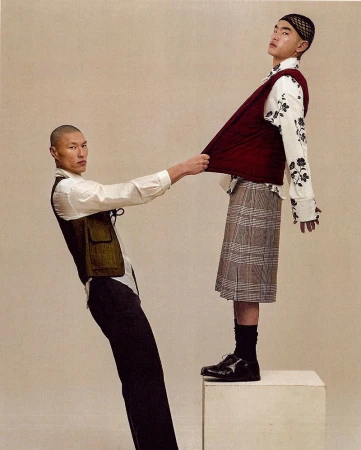 Two men in layered designer menswear and a plaid pleated skirt posing dynamically against a warm neutral studio backdrop.